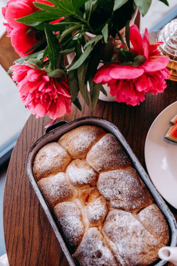 Dřevěný stůl s květinami a čerstvě upečenými buchtami posypanými moučkovým cukrem.
