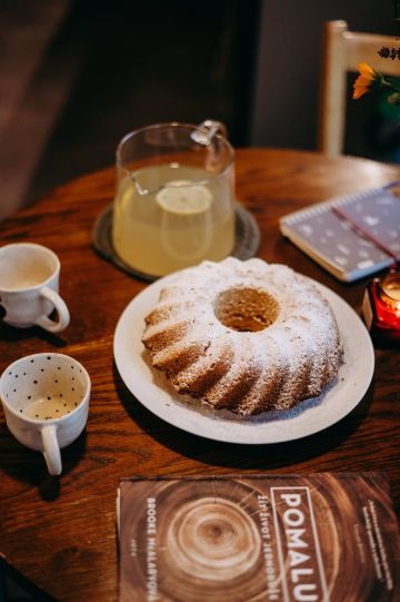 Prostřený dřevěný stůl s bábovkou posypanou moučkovým cukrem, džbánem s domácí limonádou a keramickými hrnky.