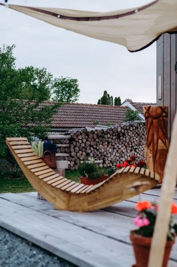 Relaxační terasa u tiny house s dřevěným lehátkem a výhledem na dřevo i louku v glampingu na statku.